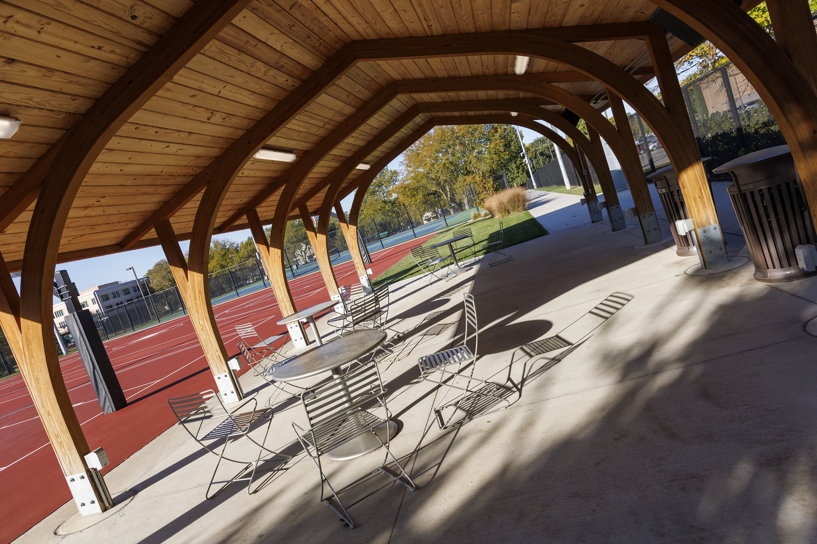 Metal table and chairs underneath a wooden covering on a cement patio with basketball court in background