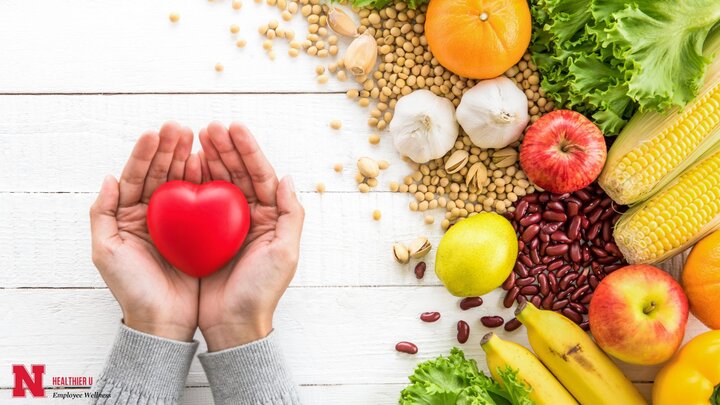 Hands holding a red heart next to a pile of veggies, grains, and fruits