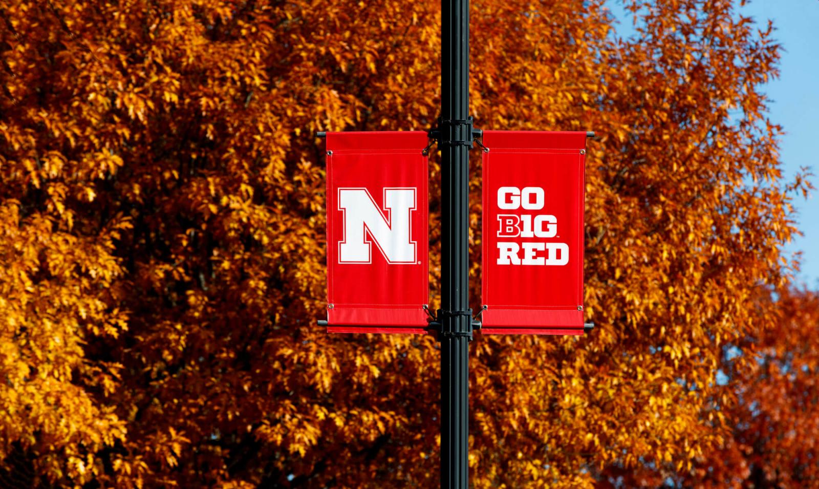 Red UNL Banners on flagpole against fall trees